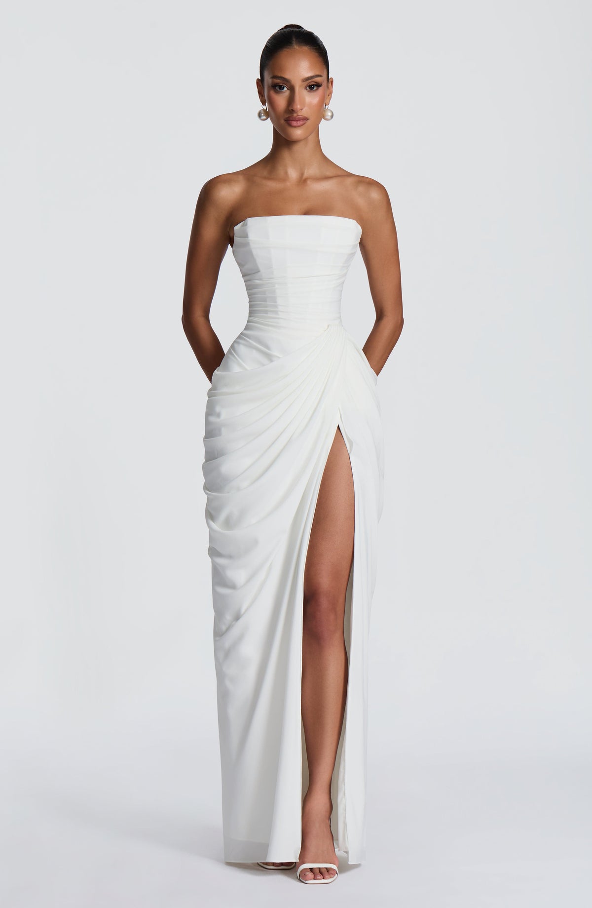Front view of the white strapless corset maxi dress with draped wrap skirt, high slit, pearl earrings and white heeled sandals. 7