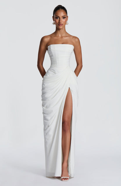 Front view of the white strapless corset maxi dress with draped wrap skirt, high slit, pearl earrings and white heeled sandals. 7