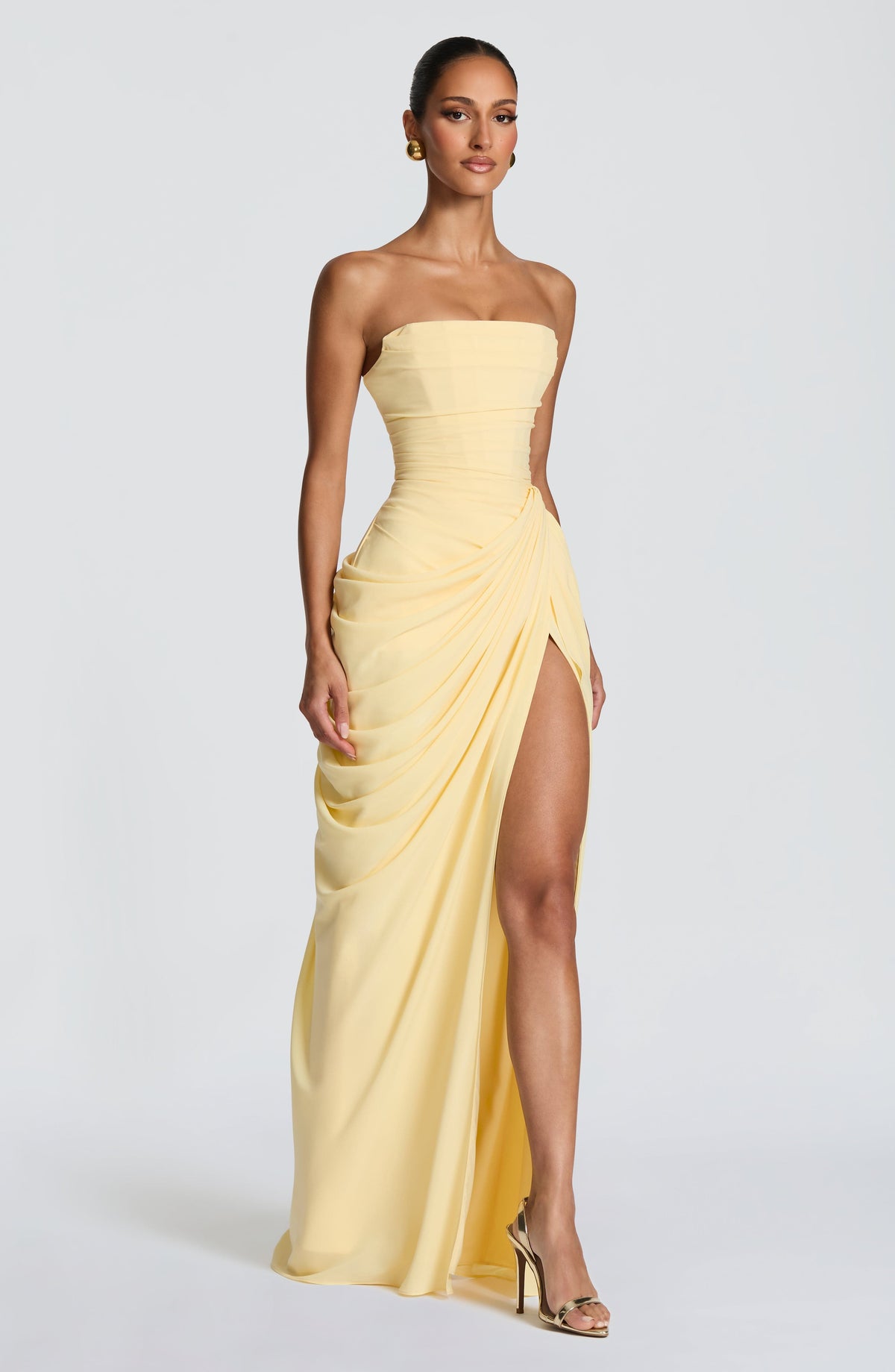 Three-quarter front view of the pale yellow strapless maxi dress, showing the draped wrap skirt and high slit with gold heeled sandals. 3