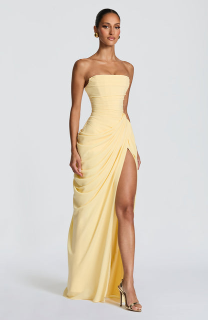 Three-quarter front view of the pale yellow strapless maxi dress, showing the draped wrap skirt and high slit with gold heeled sandals. 3