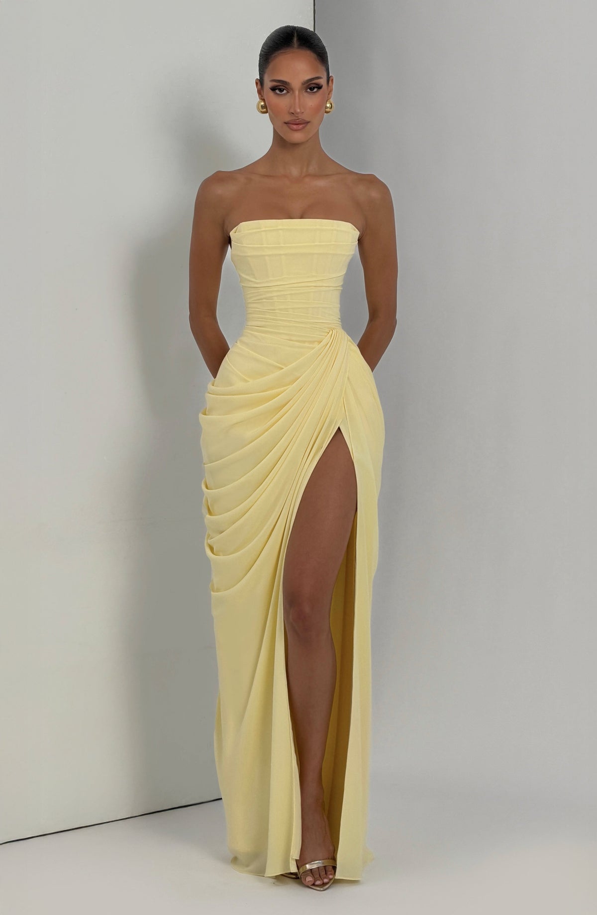 Pale yellow strapless corset maxi dress worn against a light grey studio wall, front view with high slit and gold earrings. 6