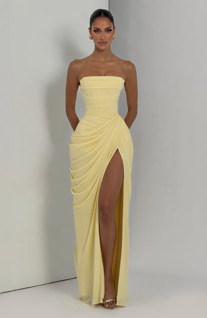 Pale yellow strapless corset maxi dress worn against a light grey studio wall, front view with high slit and gold earrings. 6
