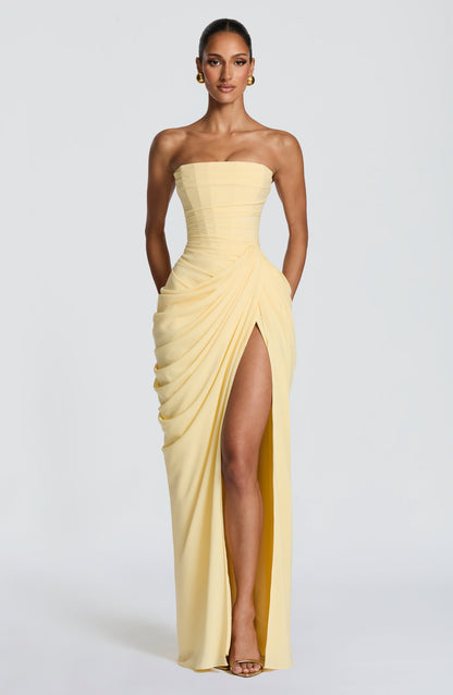 Model facing the camera in the pale yellow strapless corset maxi dress with draped skirt and high thigh slit, hands tucked in pockets. 1