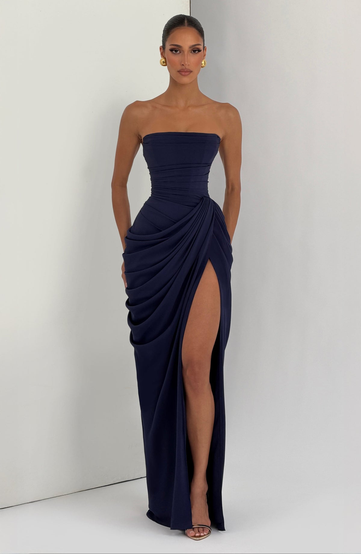 Navy strapless corset maxi dress worn against a light studio wall, front view showing the high slit and draped hip. 20