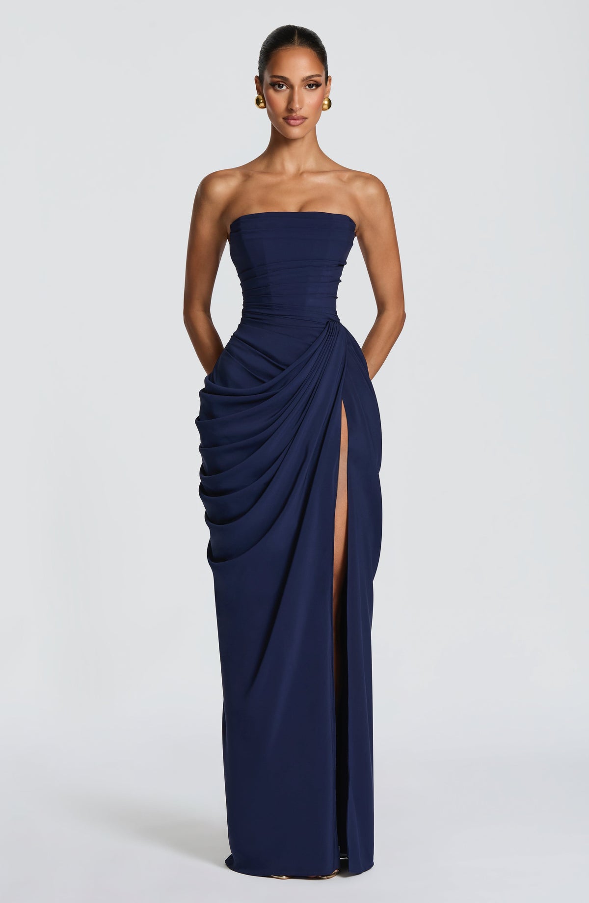 Full-length front view of the navy strapless corset maxi dress, with side drape, high slit and gold ball earrings. 19