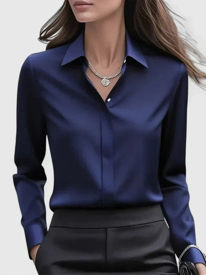 Front view of the navy satin blouse on a model, with pointed collar, concealed placket and a silver necklace, worn with black trousers. 1