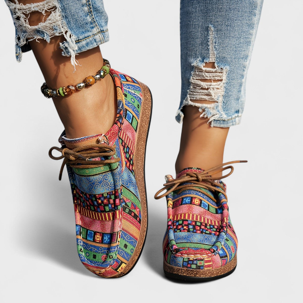 Front view of pink, blue and green patterned lace-up shoes with brown laces, worn with distressed jeans and a beaded anklet. 5