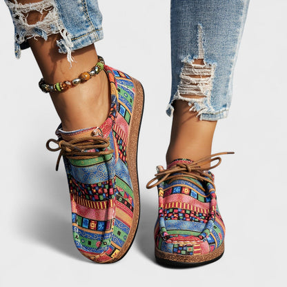 Front view of pink, blue and green patterned lace-up shoes with brown laces, worn with distressed jeans and a beaded anklet. 5