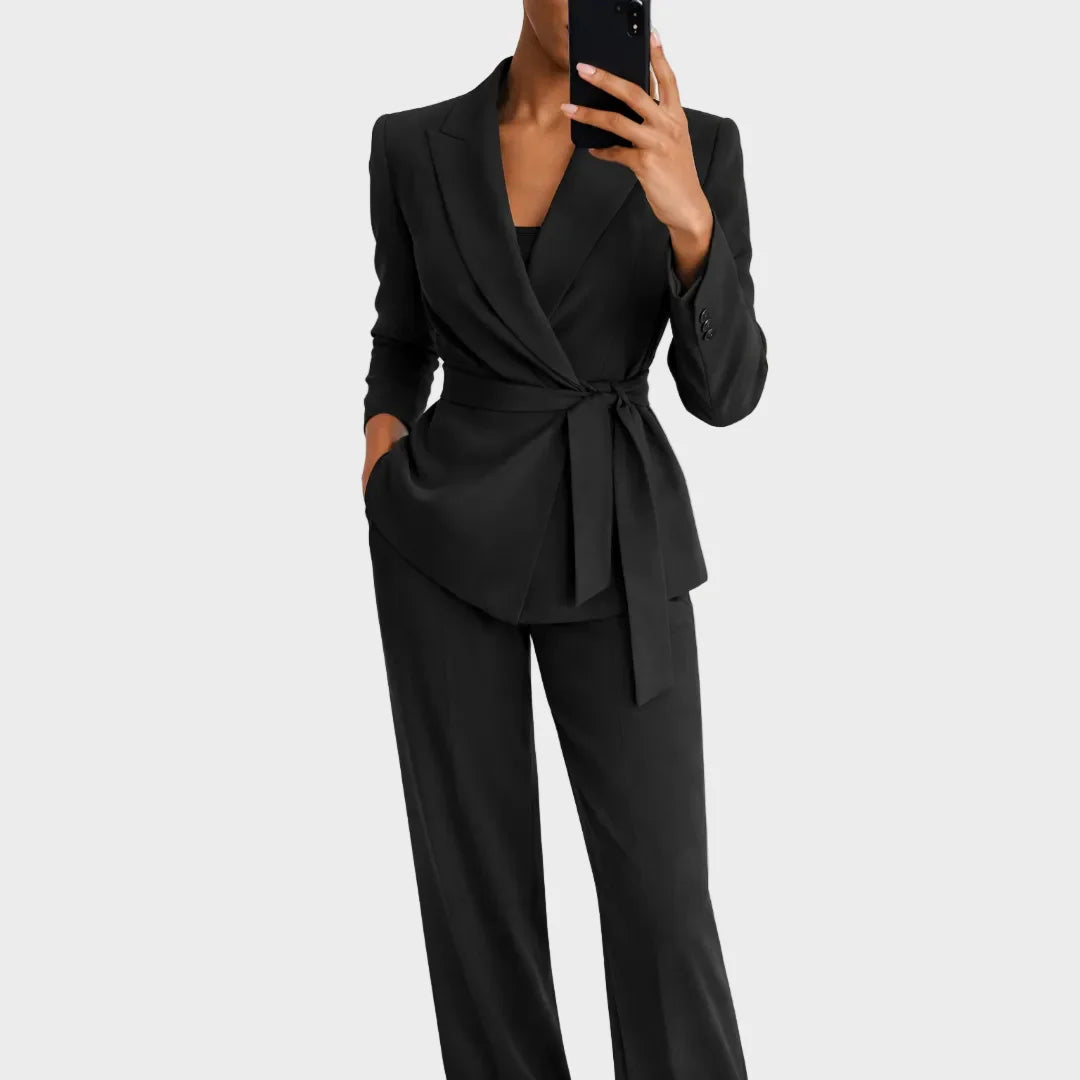 Model wearing the black version of the belted wrap blazer and matching wide-leg trousers, one hand in a pocket. 3