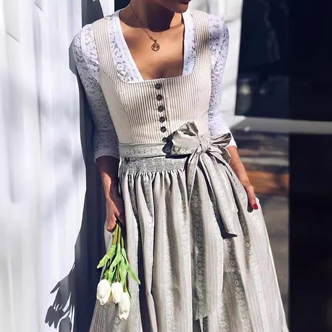 Model in the dirndl over a white lace blouse, leaning on a wall and holding white tulips, with the apron bow at the waist. 2