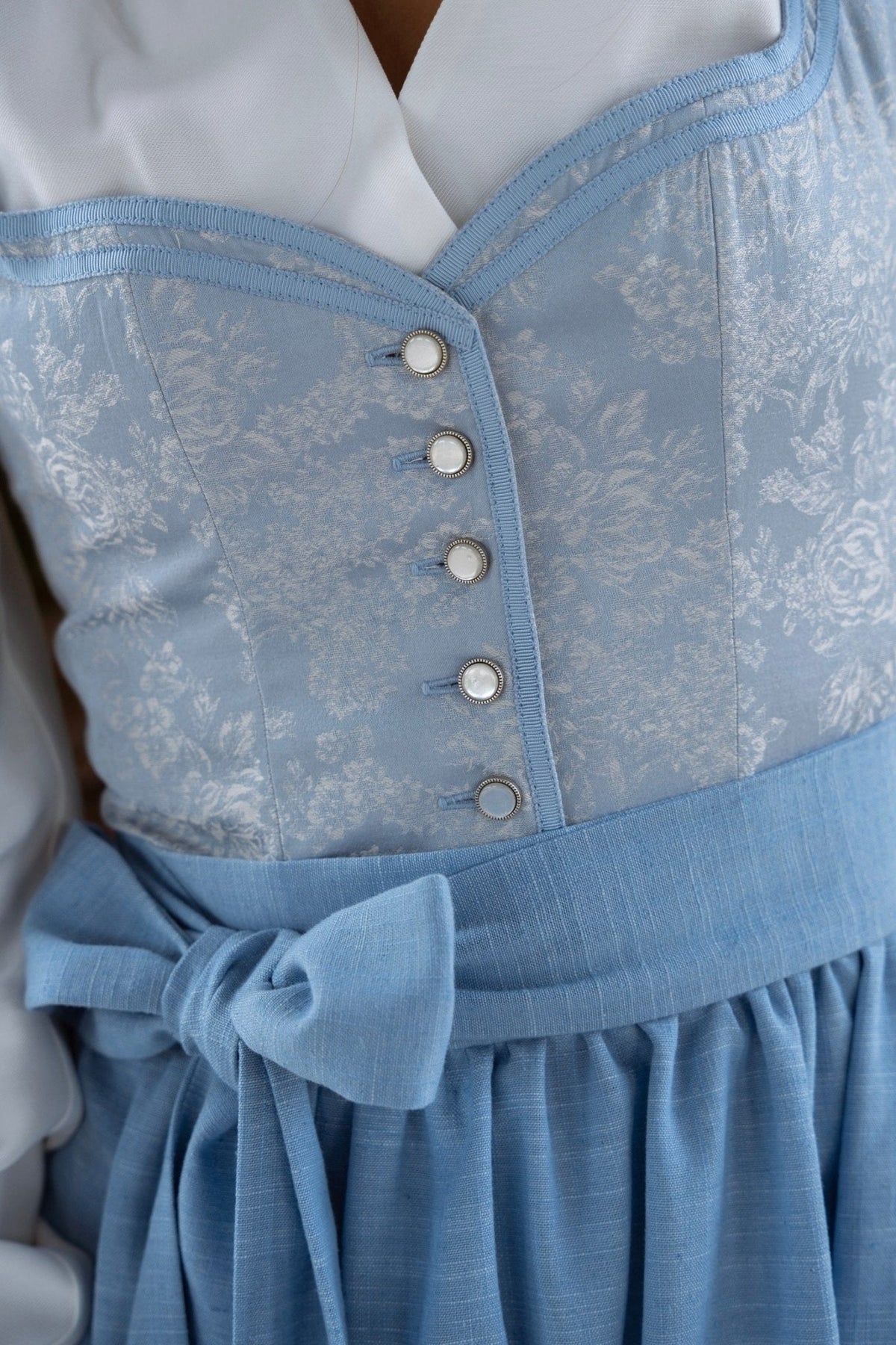 Close-up of the dirndl bodice showing five pearl-style buttons, blue trim, the floral jacquard weave and the apron tied in a bow. 2