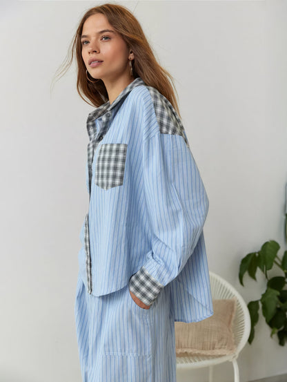 Side view of the blue-striped pyjama set with grey check collar, pocket and cuffs, worn by a model beside a chair and plant. 3