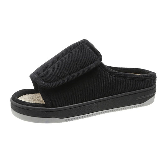 Single black corduroy open-toe slipper with a wide adjustable strap, woven insole and a grey tread sole, seen from the side. 1