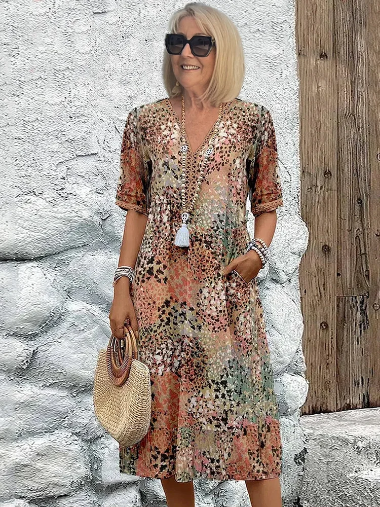Model in sunglasses wears the dress in a muted earthy speckled print of peach, green and brown against a white stone wall. 5