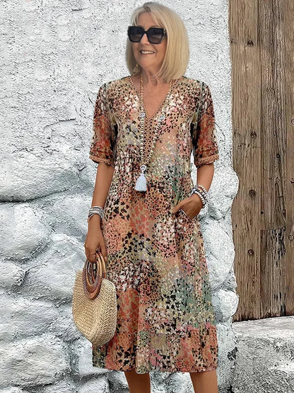 Model in sunglasses wears the dress in a muted earthy speckled print of peach, green and brown against a white stone wall. 5