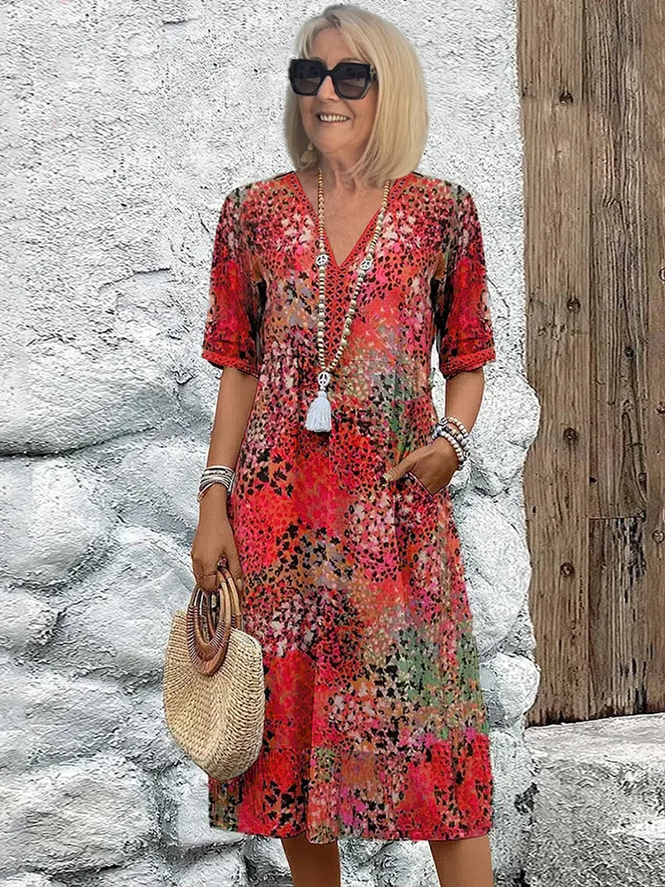 Smiling model in sunglasses wears the V-neck, half-sleeve dress in a red and pink speckled print, holding a straw bag. 1