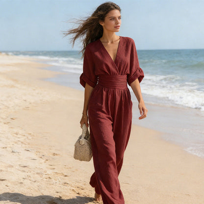 Model walking barefoot along a sandy beach in the burgundy jumpsuit, hair blowing in the breeze, carrying a woven bag. 7
