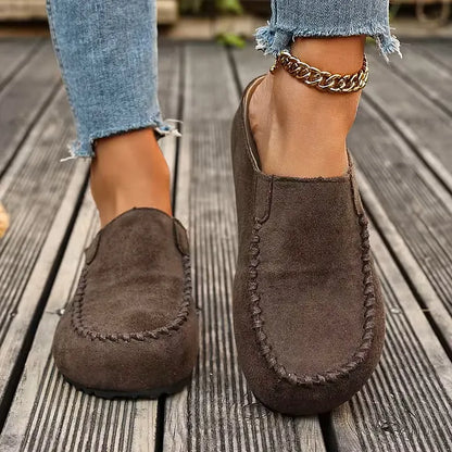 Front view of dark brown suede-look slip-ons worn on a wooden deck, showing the rounded toe and stitched seam. 4