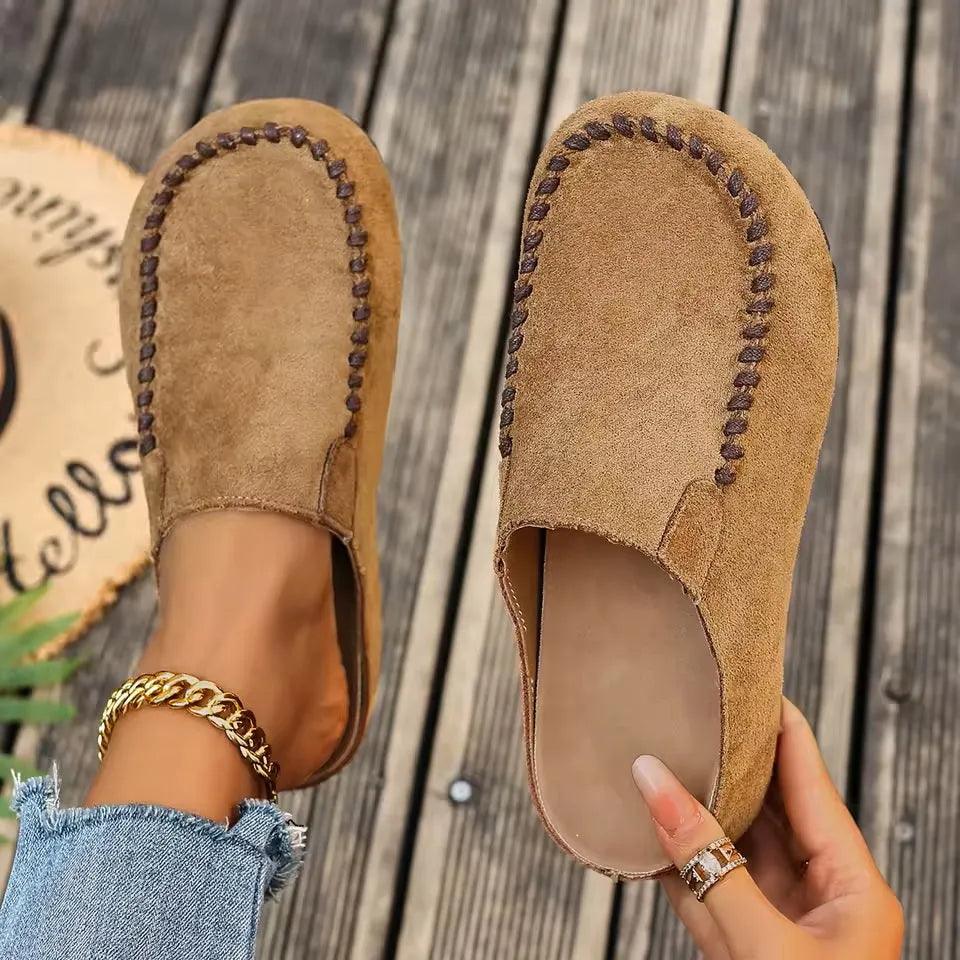Top-down view of tan suede-look slip-on clogs with brown contrast stitching, one worn and one held to show the smooth insole. 1