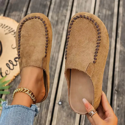 Top-down view of tan suede-look slip-on clogs with brown contrast stitching, one worn and one held to show the smooth insole. 1