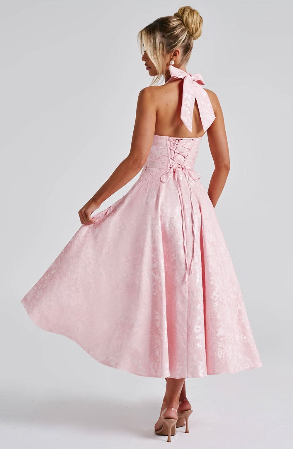Back view of the pink halter dress, with the model lifting the skirt to show the lace-up corset back and neck bow. 8