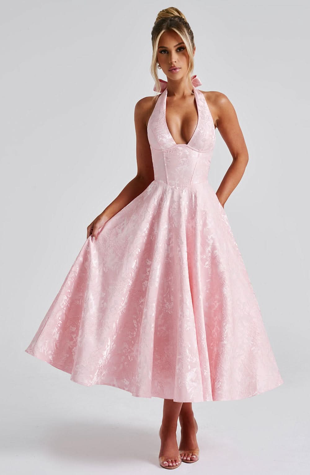 Pale pink halter midi dress on a model who holds out the full floral jacquard skirt, front view with bow at the neck. 7