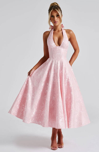Pale pink halter midi dress on a model who holds out the full floral jacquard skirt, front view with bow at the neck. 7