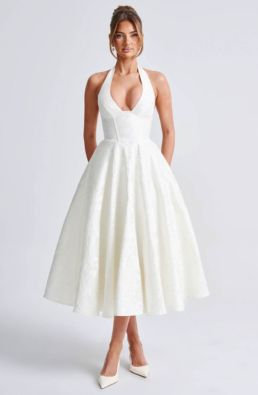 Model wearing the ivory halter midi dress facing front, with plunging neckline, structured corset bodice, full skirt and pointed white heels. 1