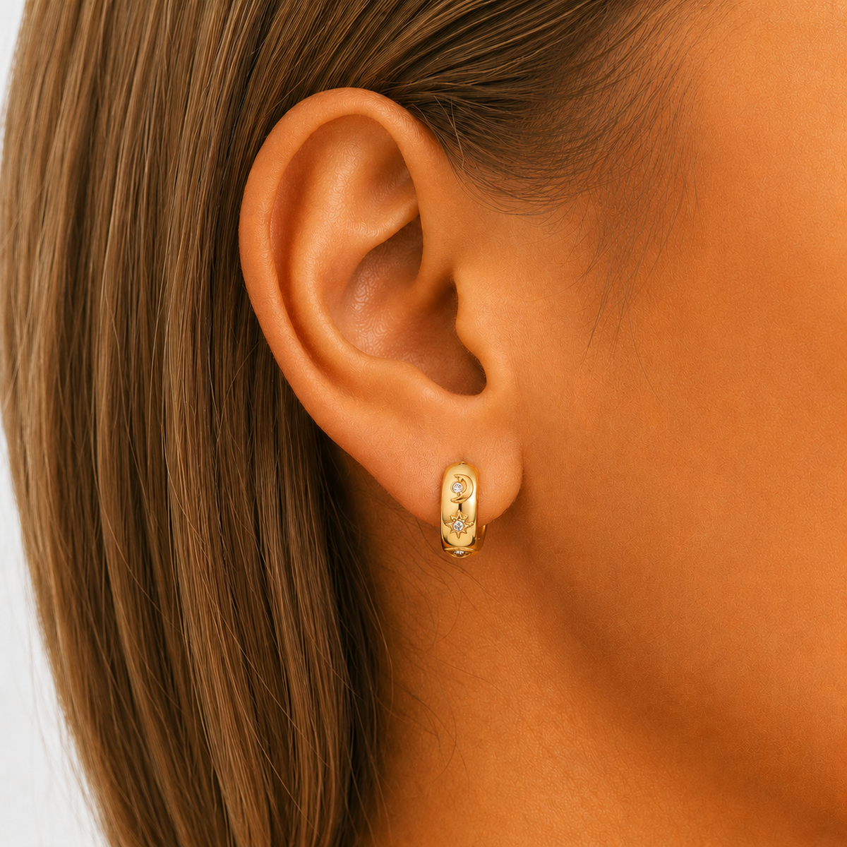 Close-up of a gold-tone huggie hoop worn in a pierced earlobe, showing the moon and star motifs with small clear stones against long brown hair. 3