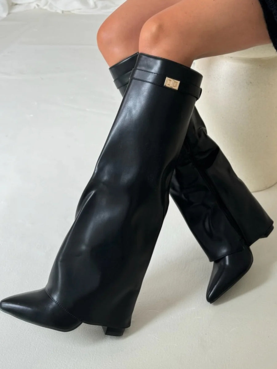 Black faux leather fold-over knee boots on a seated model, showing the smooth glossy finish and gold turn-lock strap. 15