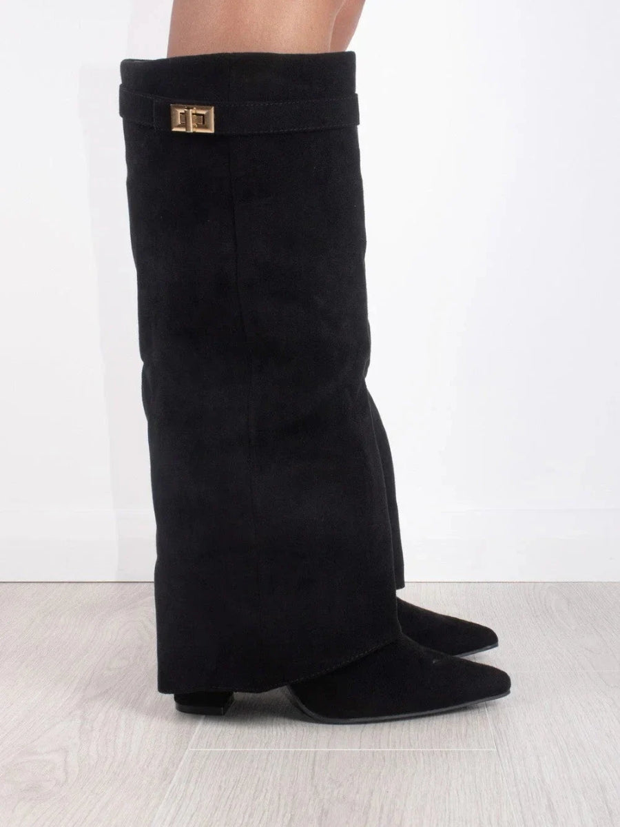 Side view of a black suede-look fold-over knee boot with pointed toe and gold turn-lock strap, worn on a grey floor. 1