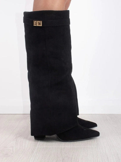 Side view of a black suede-look fold-over knee boot with pointed toe and gold turn-lock strap, worn on a grey floor. 1