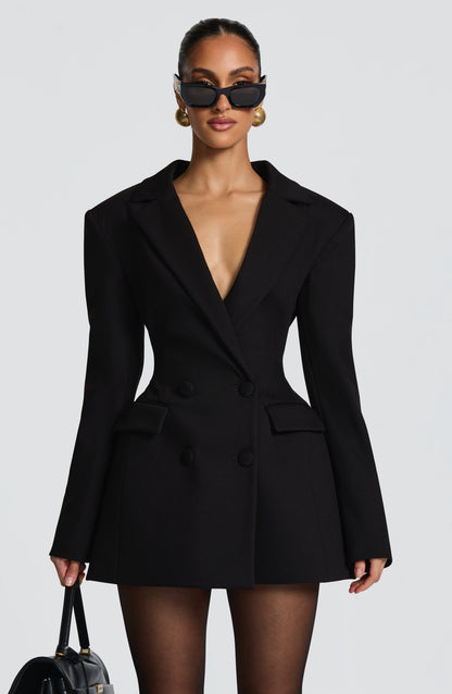 Upper-body front view of the black blazer dress with sunglasses, gold earrings, double-breasted buttons and a handbag held at the side. 10