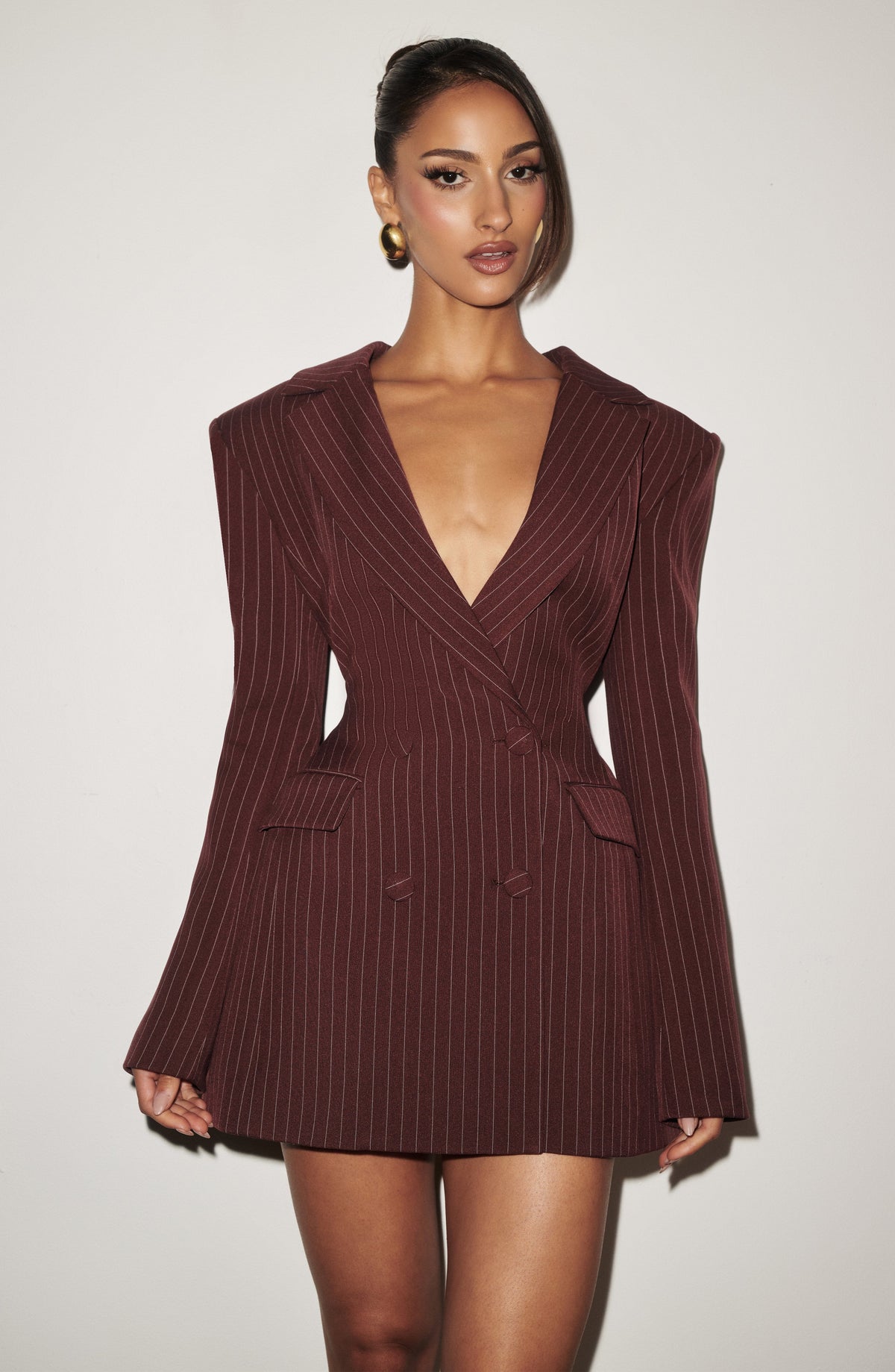 Model in a burgundy pinstriped double-breasted blazer mini dress with plunging neckline and structured shoulders, photographed against a plain white wall. 21