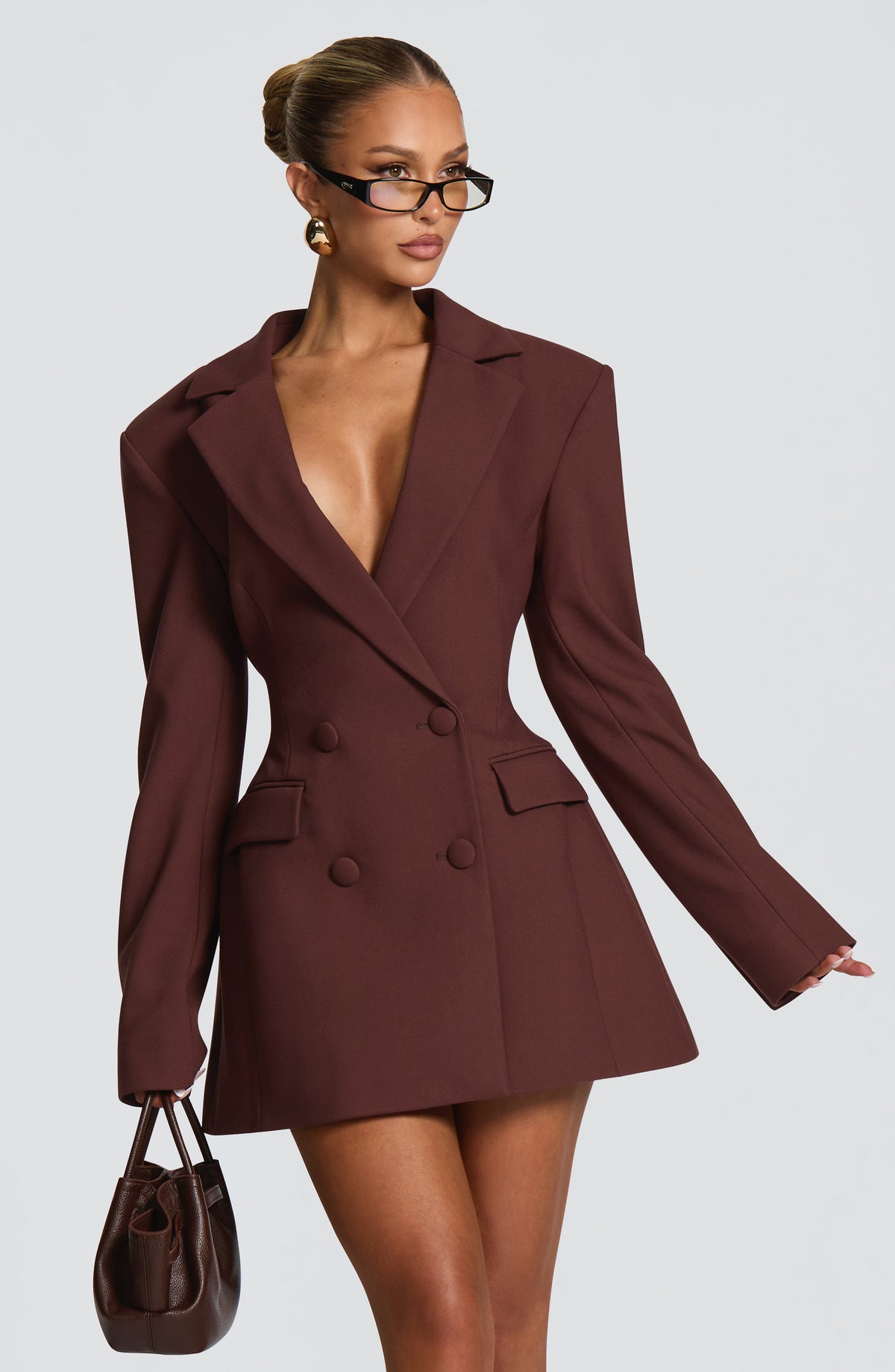 Brown double-breasted blazer dress, three-quarter view on a model holding a small brown leather handbag, showing the nipped waist and flared skirt. 2