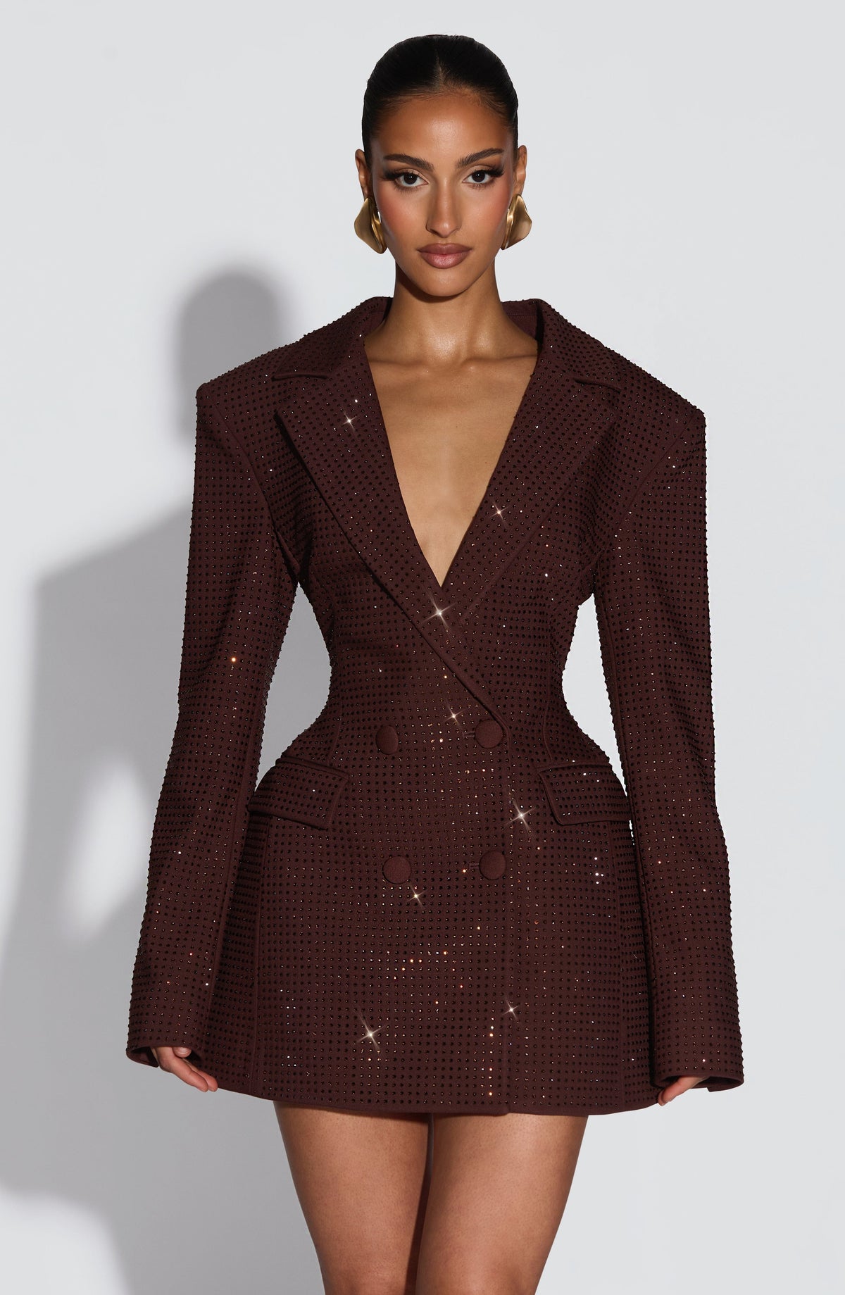 Front view of a dark brown blazer dress covered in sparkling crystal embellishment, with a plunging neckline and gold sculptural earrings. 19