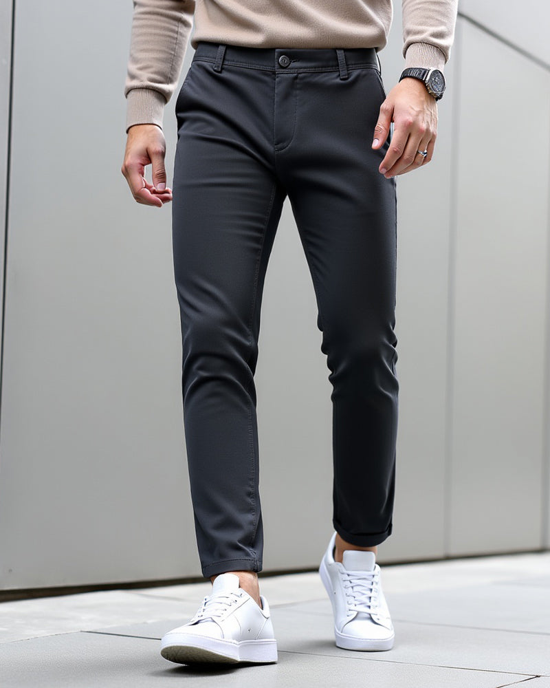 Dark charcoal chinos with turned-up hems worn with a beige knit jumper, watch and white trainers against a grey wall. 9