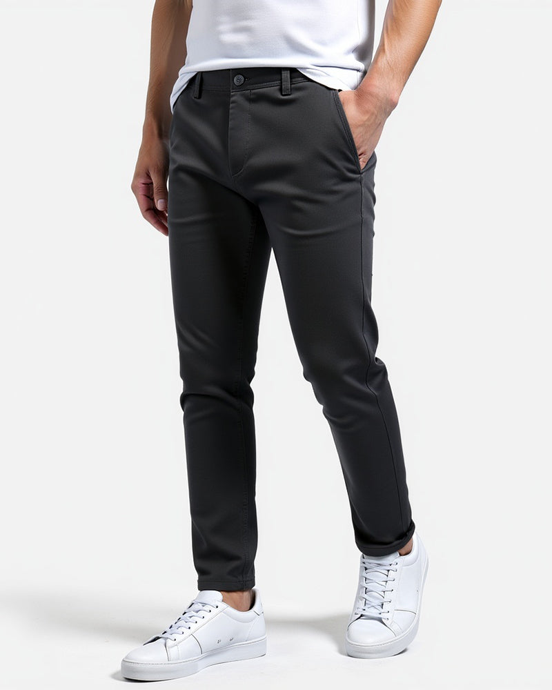 Dark charcoal chinos worn by a model with a white t-shirt and white trainers, one hand in the pocket, angled front view. 6
