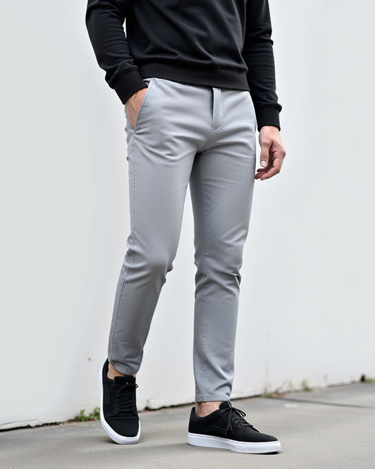 Light grey chinos on a model in a black sweatshirt and black trainers, standing against a white wall outdoors. 2