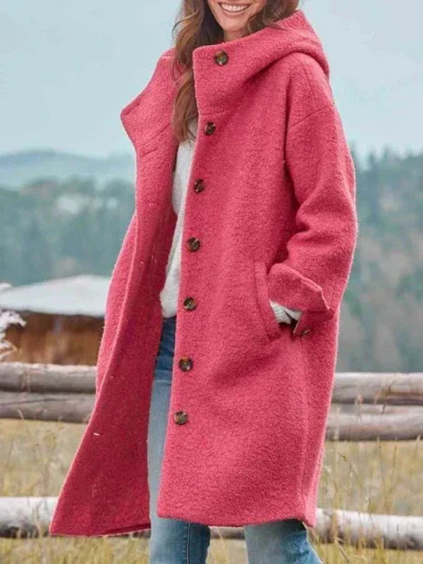 Smiling woman wearing the hooded, button-front coat in coral pink with a textured finish, one hand in the side pocket, by a rustic fence. 1