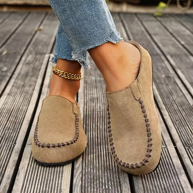 Front three-quarter view of beige slip-on clogs worn with cropped frayed jeans, showing the brown stitching and open back. 5