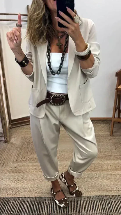 Woman wearing the light stone-coloured blazer with patch pockets and matching rolled-hem trousers, styled with a chunky chain necklace and wide belt. 2