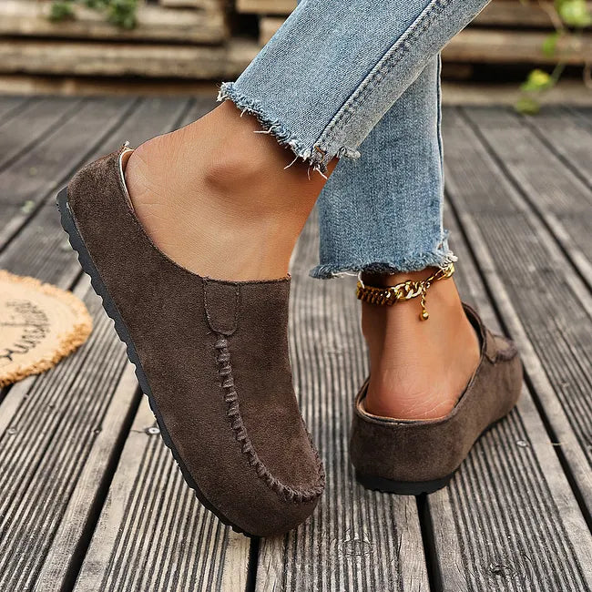 Side and back view of dark brown slip-ons worn mid-step, showing the open heel and thin black flat sole. 6