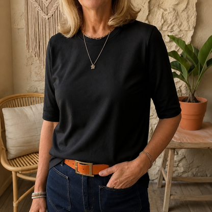 Woman wearing the round-neck half-sleeve t-shirt in black, tucked into dark jeans with a tan belt, in a cosy room with a rattan chair. 7