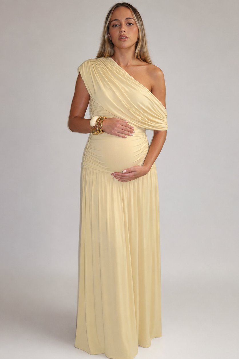 Front view of a pregnant model in the pale yellow one-shoulder maxi dress with draped bodice, ruched waist and gold bangles. 1