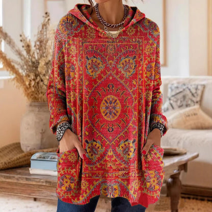 Wider shot of a model wearing the red hooded tunic with a rug-style medallion print, hands in the front pockets, in a living room. 1