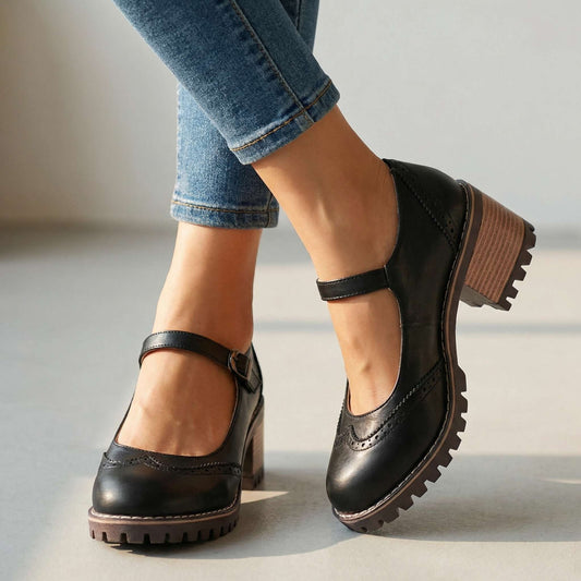 Black brogue-detail shoes with ankle strap and block heel, worn with rolled blue jeans against a plain pale studio floor. 1