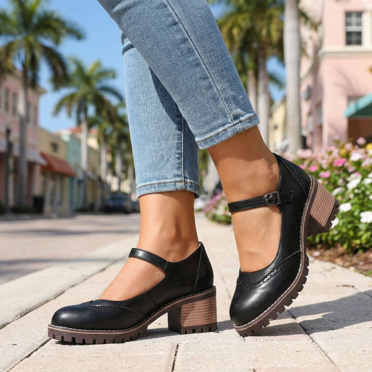 Black pair worn on a pavement beside flowers, side view with buckle strap, brogue perforations and stacked heel 7