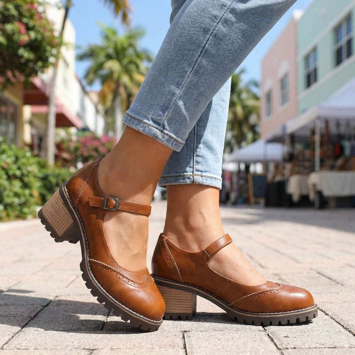 Tan-brown pair worn on a sunny paved street lined with palm trees, showing the buckle strap and brogue toe detailing. 5