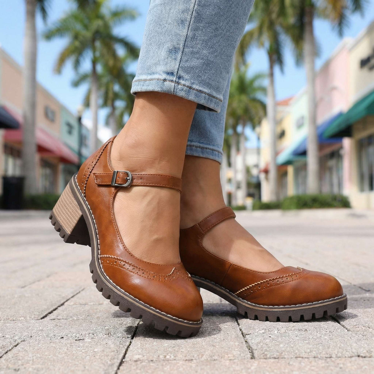 Close low-angle view of tan-brown shoes on paving, highlighting the buckle, contrast stitching around the brogue toe and stacked heel. 8
